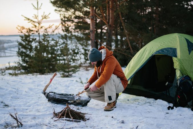 A man sitting near his tent prepares wood wool with a knife for starting a fire in a winter forest at sunset.
